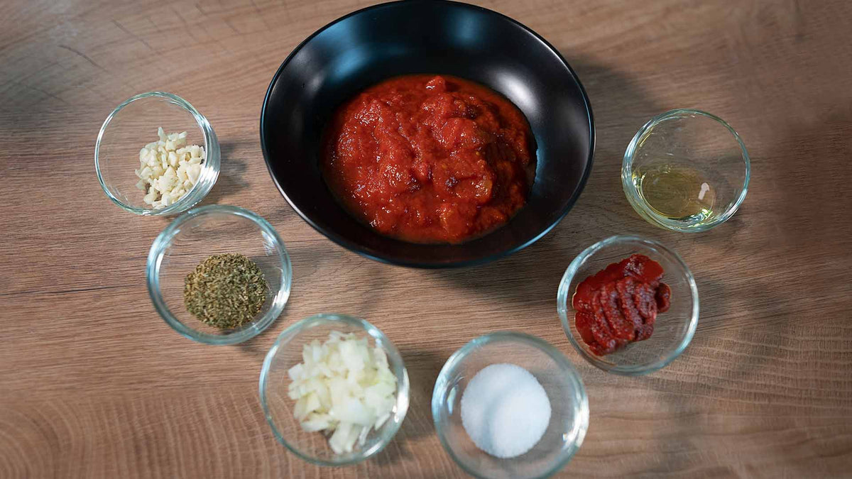Eine schwarze Schüssel mit Tomatensoße ist von kleinen Glasschalen umgeben, die gehackten Knoblauch, getrocknete Kräuter, gehackte Zwiebeln, Tomatenmark, Zucker, Öl und Salz enthalten, alles auf einer Holzoberfläche angeordnet.