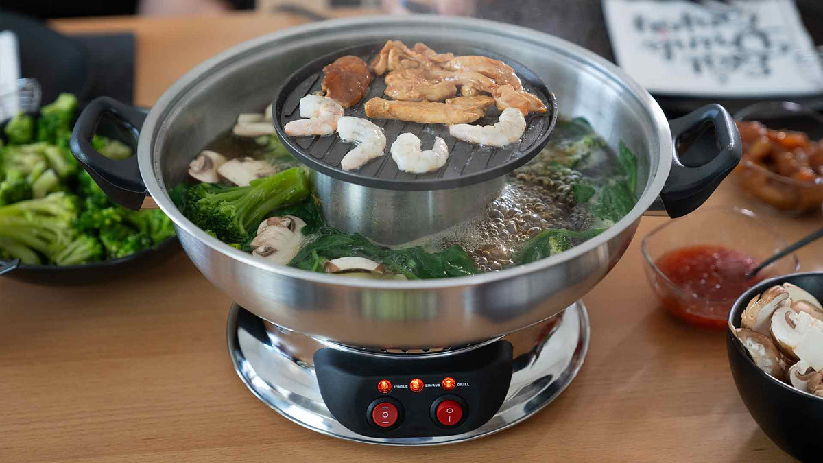 Ein Eintopf mit kochender Brühe enthält Gemüse und Pilze, während darüber auf einem Grill Garnelen, Fleisch und Tofu garen. Teller mit Gemüse und eine Schüssel mit Soße stehen daneben auf dem Tisch.