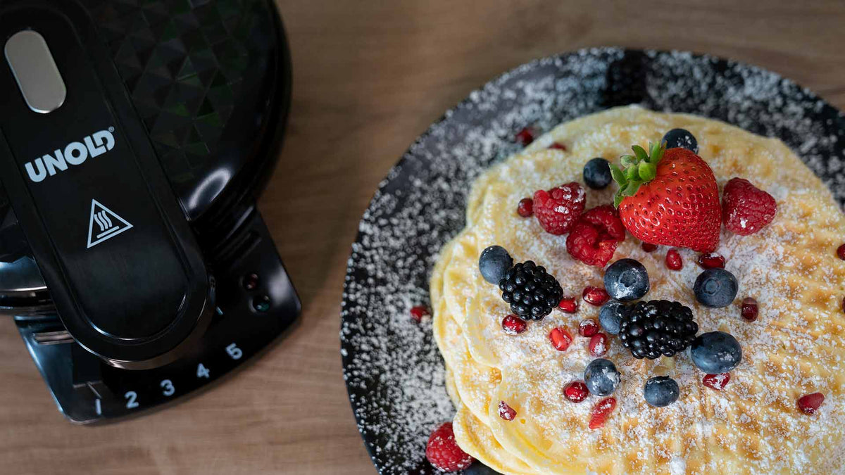 Ein Stapel Pfannkuchen mit Puderzucker, Erdbeeren, Himbeeren, Blaubeeren, Brombeeren und Granatapfelkernen auf einem schwarzen Teller, neben einem schwarzen UNOLD-Waffeleisen auf einer Holzoberfläche.