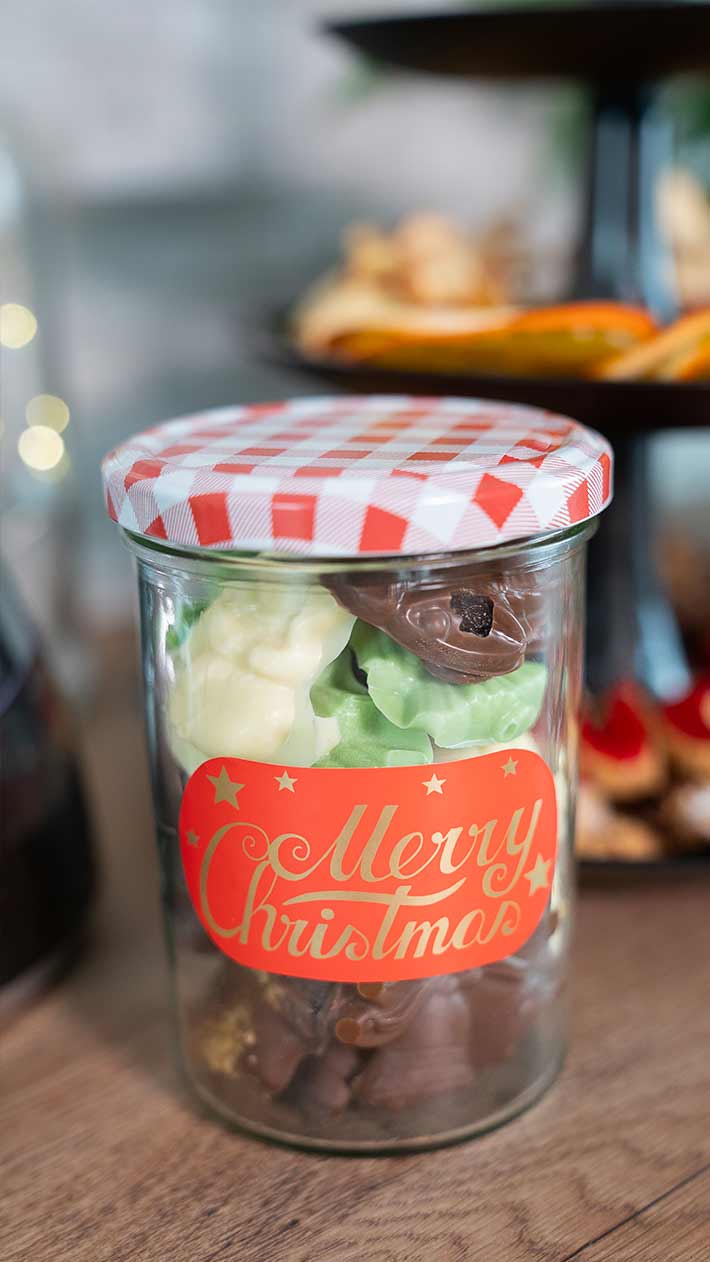 Ein Glasgefäß mit rot-weiß kariertem Deckel und der Aufschrift „Frohe Weihnachten“, gefüllt mit verschiedenen Pralinen und Süßigkeiten, steht auf einer Holzoberfläche neben einem Teller mit Leckereien im Hintergrund.
