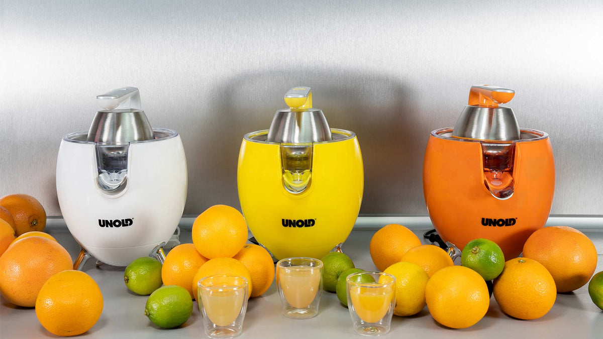 Drei elektrische Zitruspressen in Weiß, Gelb und Orange stehen auf einer Theke. Davor stehen frische Orangen, Limetten und Gläser mit Saft.