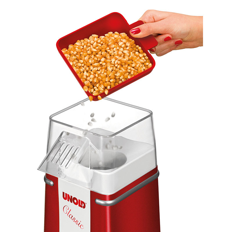 Eine Hand schüttet gelbe Popcornkörner aus einer roten Schaufel in die obere Öffnung eines Unold Popcornmaker Classic, der ein rot-weißes Design aufweist.